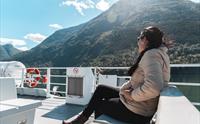 Passasjer sit på utedekket i solskinn under fjordcruise på Hardangerfjorden med grøne fjellsider rundt.