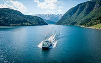 Fjordcruisebåten på Hardangerfjorden sett frå drone forfra, med blått vatn og snødekte fjell på begge sider.