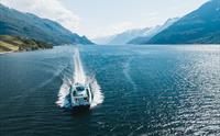 Fjordcruisebåten frå drone over Hardangerfjorden med solrefleksar i vatnet og frukttre i lia på eine sida.