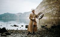 En kvinne utkledd som en viking, som står ved fjorden og spiller en vikingtromme.