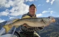 En mann smiler mens han holder en stor fisk ombord på en båt med fjell i bakgrunnen.