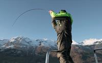 En fisker i grønn jakke kjemper med en fiskestang ombord på en båt, med snødekte fjell i bakgrunnen.