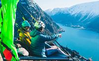 To personar sit på ein portaledge ved fjorden i Hardanger, med majestetiske fjell i bakgrunnen. Ein unik overnattingoppleving i fjellveggen.
