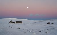 Noen fjellhytter i vinterlandskapet under en rosa himmel med fullmåne i bakgrunnen.