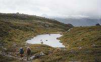 To turgåere som går forbi et lite fjellvann, med fjell i bakgrunnen.