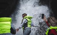 En kvinne foran i båten blir «dusjet» av en foss, andre passasjerer tar bilder med telefonene sine.