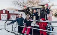 Barn i vinterklær og nisseluer som nyter en snøfylt dag, smilende og poserende ved siden av et snøkjøretøy i en festlig, gårdslignende omgivelse.