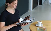 Ei ung kvinne sit ved eit bord og leser i ein bok, med ein kopp kaffi og eit kakestykke foran seg.