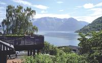 Utsikt ut over fjord og fjell. I forgrunnen er det grønne trær og en altan hvor det sitter tre mennesker.