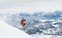 En person med rød jakke hopper utfor en skråning på ski. Utsikt til fjord og snødekket landskap i bakgrunnen.