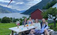 Glade gjester nyt sommarkvelden ved eit utandørs piknikbord med utsikt over fjorden på Jaastad Gjestetun.