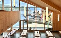 Stort lokale med stor takhøyde med store vinduer med utsikt over fjorden. Mange langbord står oppstilt og det er god plass til å sitte. Utsikt over båtkai og fjorden.