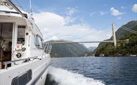 Pilagutt på veg under ei imponerande bru i Hardangerfjorden, omkransa av fjell og blå himmel.