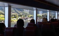 Reisande nyt fjordutsikta frå innsida av Hardangerfjordekspressen, med landsbyen og fjellandskapet i bakgrunnen.