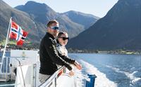 Eit smilande par på Hardangerfjordekspressen med norsk flagg og naturskjønne omgjevnadar i bakgrunnen.