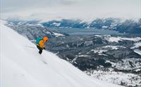 En oransje og gulkledd person står på ski ned en bratt fjellside med utsikt til snødekket landskap på topptur i Hardanger.