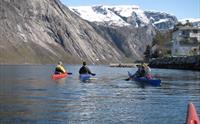 Kajakkpadlere i Eidfjord med snødekte fjell i bakgrunnen og et tradisjonelt norsk hus ved siden av fjorden.