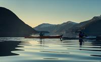 Kajakkpadlere på stille vann i Hardangerfjorden ved solnedgang, med fjell silhuetter og refleksjoner i vannet.